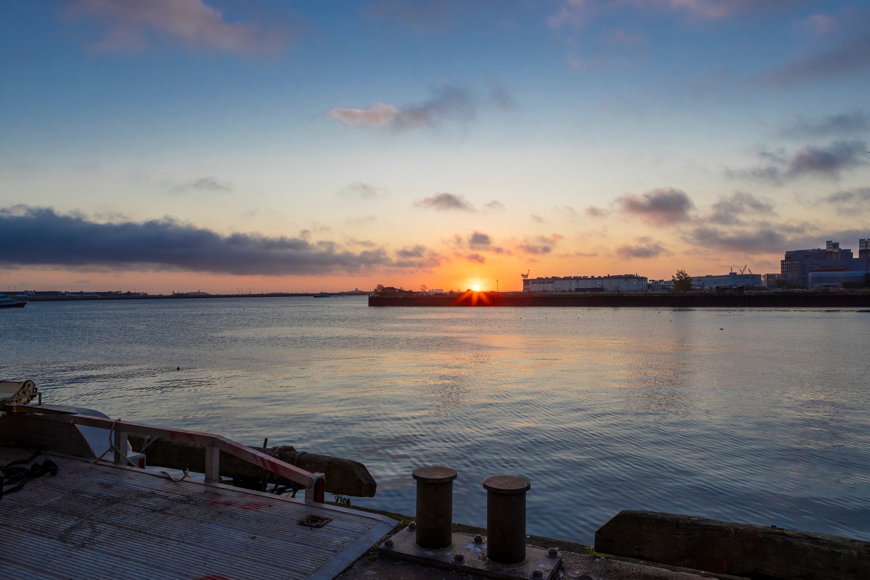 A scenic sunset over a calm body of water with a pier in the foreground and buildings on the horizon under a blue and orange sky.